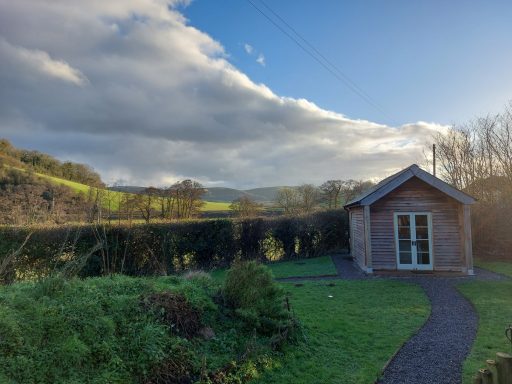 A wooden cabin surrounded by greenery and hills under a cloudy sky.