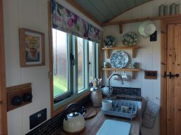 Cosy kitchen with wooden shelves, a kettle, and floral curtains and a view outside.