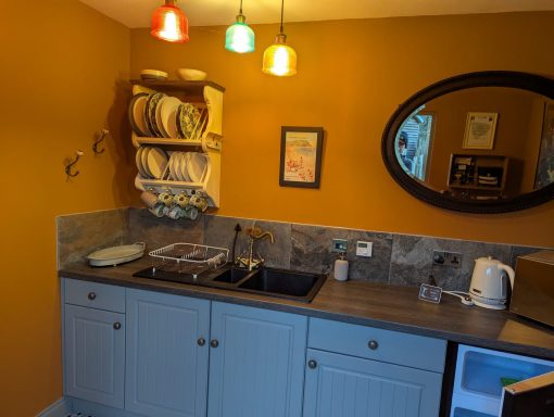 Cosy kitchen with orange walls, shelves of dishes, and colourful pendant lights.